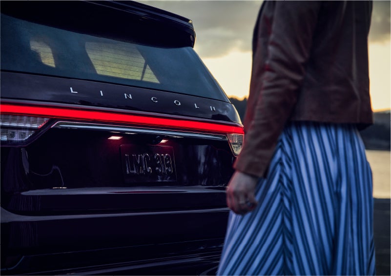 A person is shown near the rear of a 2023 Lincoln Aviator® SUV as the Lincoln Embrace illuminates the rear lights | Parks Lincoln of Gainesville in Gainesville FL